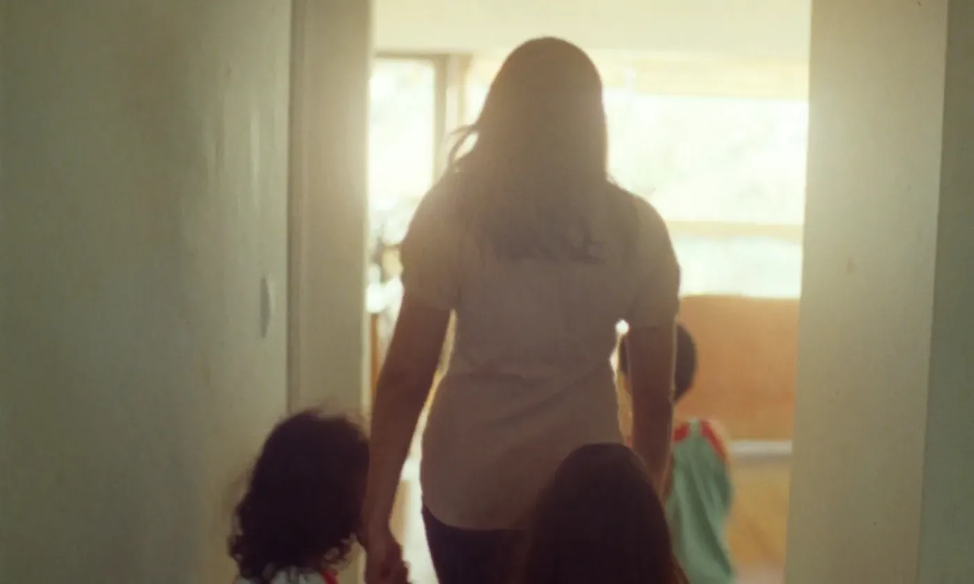 A mother walking hand in hand with her children through a sunlit home, seen from behind